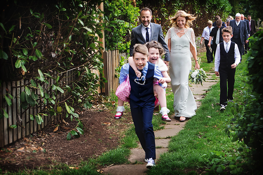 bride and groom walking from church with guests children giving piggy back captured by East Midlands Wedding Photographer